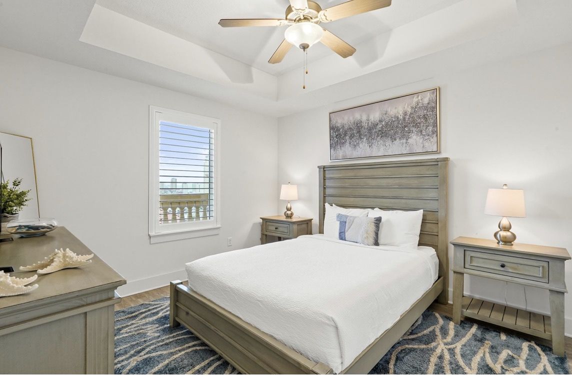 Neutral-toned bedroom with gray wooden bed, white bedding, two matching nightstands and lamps, ceiling fan, and abstract art above headboard