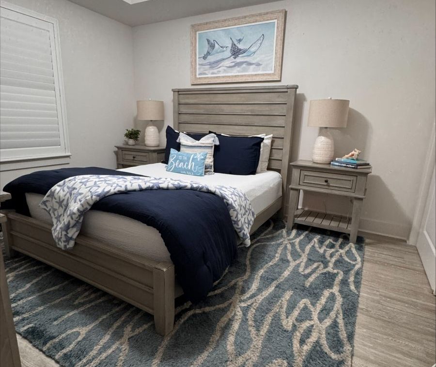 Coastal-themed bedroom with a gray wooden bed, navy and white bedding, and matching nightstands with lamps on both sides.